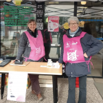 Deux bénévoles de Ma P'tite Echoppe en tablier rose des Restos du coeur attendant les dons devant l'épicerie.